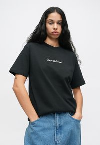 T-shirt de algodão preta com mangas curtas, apresentando um logo branco da "New Balance" no peito, combinada com calções de ganga de tom azul claro.