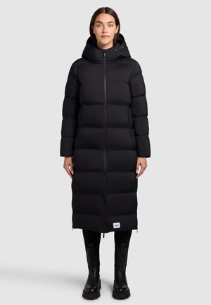 Vrouw gekleed in een lange zwarte pufferjas met capuchon, met een ritssluiting aan de voorkant, zwarte leggings en zwarte laarzen, staand tegen een effen lichte achtergrond.