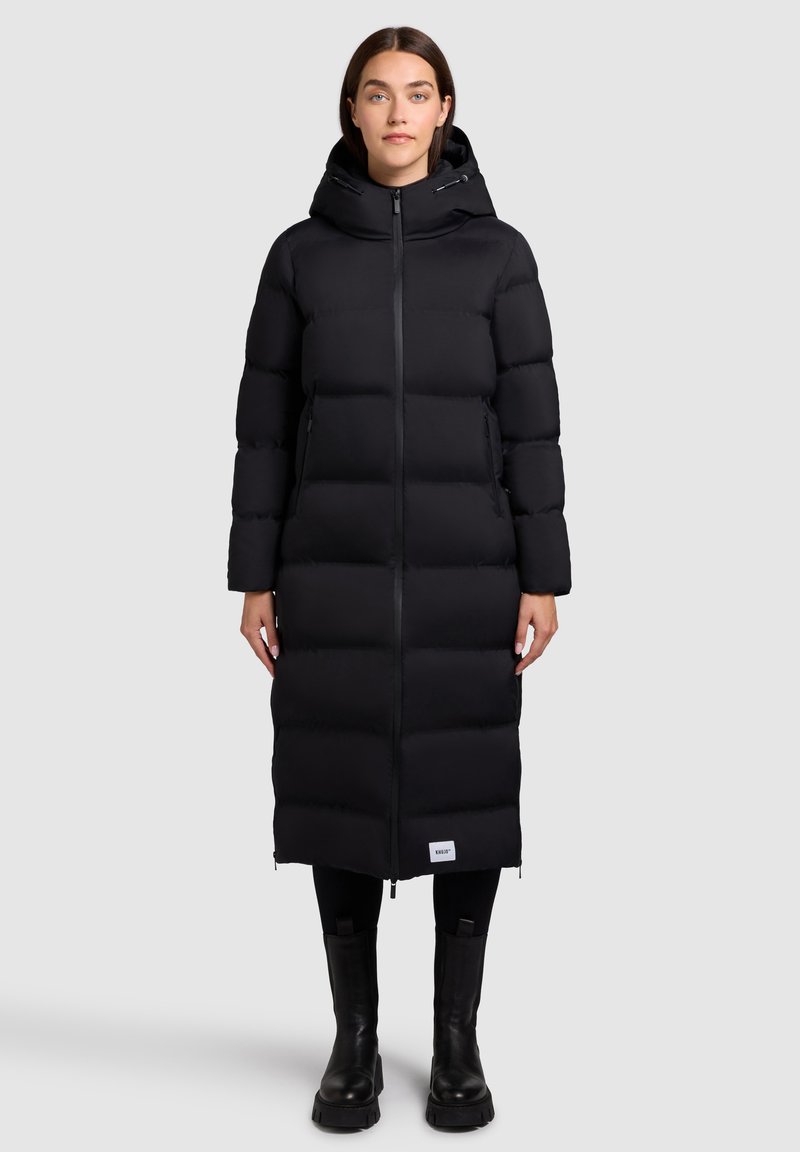 Vrouw gekleed in een lange zwarte pufferjas met capuchon, met een ritssluiting aan de voorkant, zwarte leggings en zwarte laarzen, staand tegen een effen lichte achtergrond.