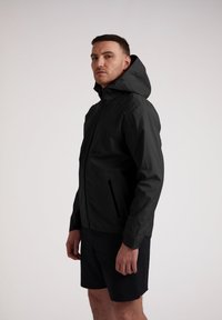 Mand i sort hættejakke og sorte shorts står med ansigtet vendt fremad mod en ensfarvet hvid baggrund.
