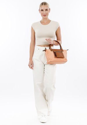 Tan handbag with brown accents, structured design, dual handles, zipper closure, and a front pocket detail. Model wears light-colored casual attire.