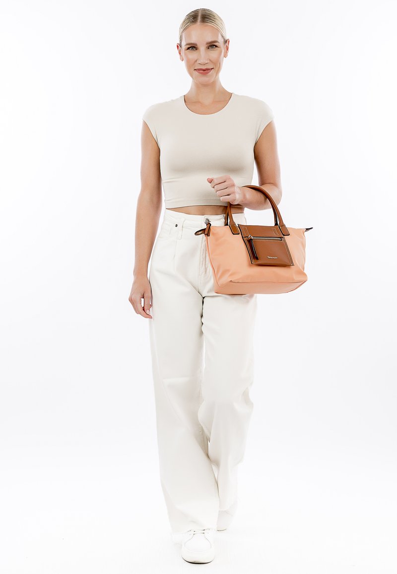 Borsa tan con dettagli marroni, design strutturato, doppi manici, chiusura a zip e tasca frontale. Il modello indossa un abbigliamento informale di colore chiaro.