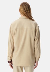 Beige overhemden met lange mouwen in een relaxed fit, voorzien van een rugnaad detail en knoopmanchetten. Soepele textuur met minimalistisch ontwerp.
