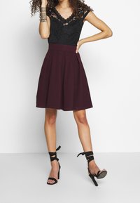 Top in pizzo nero con scollatura a V profonda, abbinato a una gonna A-line plum. La modella indossa tacchi alti neri con cinturini. Texture morbide e motivi dettagliati.