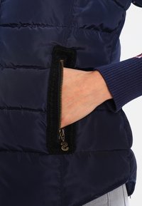 Main partiellement dans la poche ouverte d'une veste matelassée bleu marine, avec une fermeture éclair dorée visible et un poignet côtelé bleu marine sur la manche.