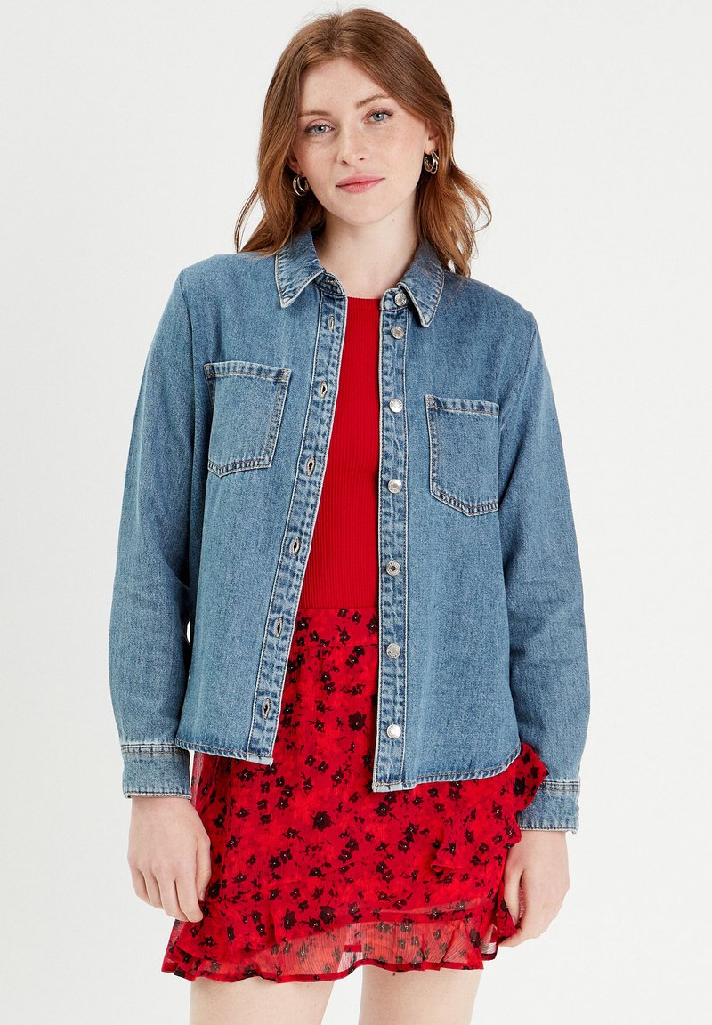 Camicia di jeans azzurro chiaro con maniche lunghe, chiusura a bottoni e due tasche sul petto, indossata sopra un top rosso a coste e una gonna floreale a strati.