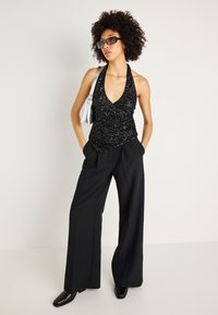 Top halter preto com lantejoulas e decote em V profundo, combinado com calças largas de cintura alta pretas e sapatos pretos de bico fino. Acento de bolsa prateada.