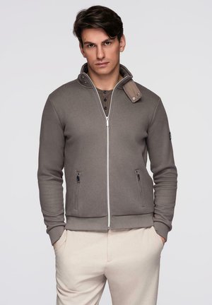 Młody mężczyzna w szarej kurtce zapinanej na zamek, nałożonej na zapiętą koszulę, z rękami w kieszeniach kremowych spodni, patrzący prosto przed siebie.