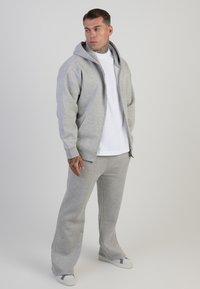 Jasnoszara bluza z kapturem, z zamkiem z przodu i dużymi kieszeniami, zestawiona z pasującymi spodniami dresowymi. Gładka tekstura i luźny krój.