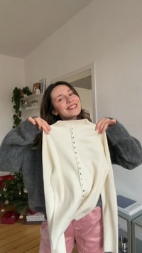 Pulover crem cu dungi și guler înalt, având un rând de nasturi argintii pe față, ținut în mâini. Purtat dedesubt un strat exterior gri cu textură.