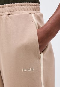 Χέρι με σκούρο χρώμα δέρματος σε τσέπη μπεζ βαμβακερού παντελονιού φούτερ με ελαστική μέση και λογότυπο "GUESS" κοντά στη πλευρική ραφή.