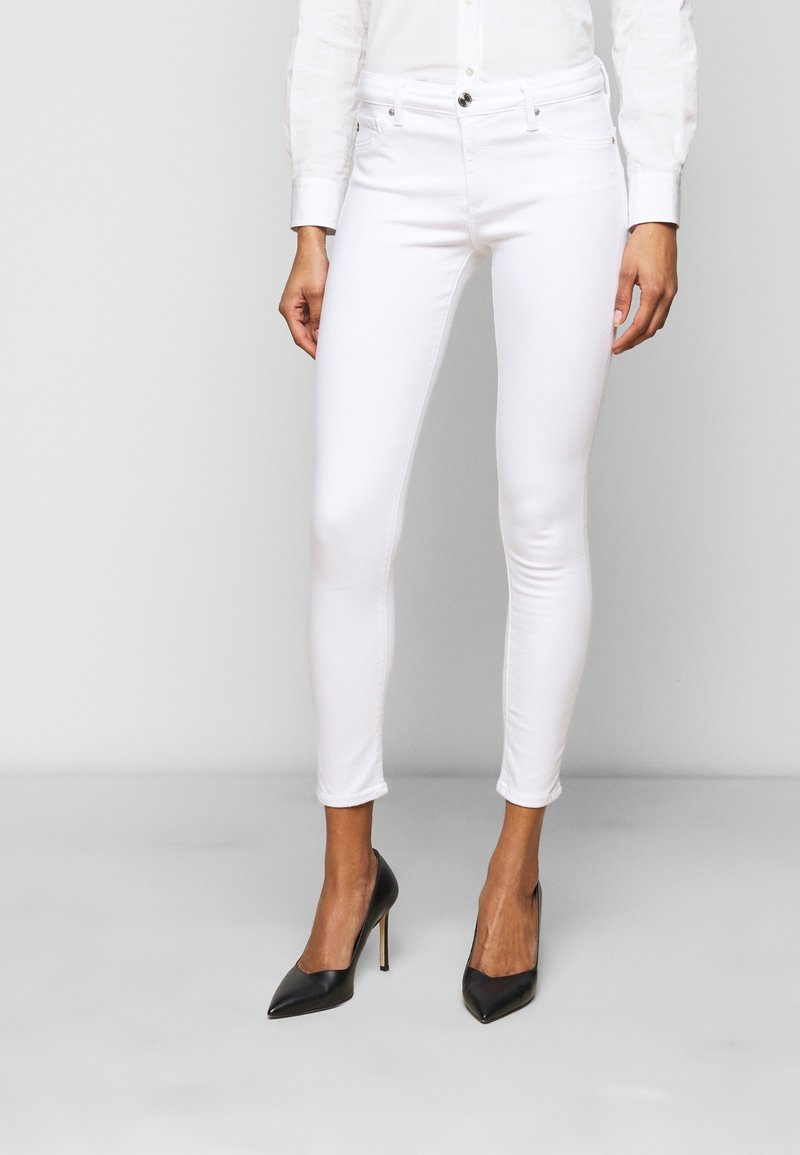 Jeans skinny bianchi in cotone elasticizzato, con un classico design a cinque tasche, chiusura frontale con bottone e una vestibilità slanciata, abbinati a tacchi neri.