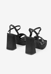 Sandalias negras de tacón alto con un tacón cuadrado grueso, diseño de punta abierta y correas entrelazadas. Textura suave y mate con hebilla ajustable.