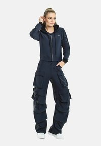 Mörkblå tvådelad outfit som består av en zip-up huva jacka och cargo-stil byxor med flera fickor och rynkiga tygdetaljer.