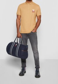 Polo beige avec logo, jeans gris et sac de sport bleu marine avec des rayures blanches et des accents marron. Bottines noires avec détails en motif.