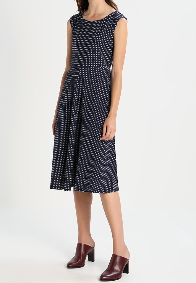 Femme portant une robe midi sans manches bleu marine à pois blancs et des mules marron à talons sur un fond uni.