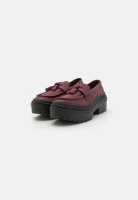 Burgundy slip-on shoes with a chunky black sole, featuring tassel accents and a smooth leather texture. Rounded shape and soft edges.