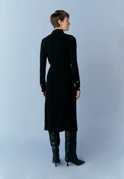 Long black dress with a slim fit, long sleeves, and a high collar; features a gathered waist and decorative button accents on cuffs.