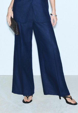 Osoba ubrana w ciemnoniebieskie spodnie o szerokich nogawkach i czarne sandały na obcasie, trzymająca czarną teksturowaną kopertówkę z srebrnymi bransoletkami na nadgarstku.