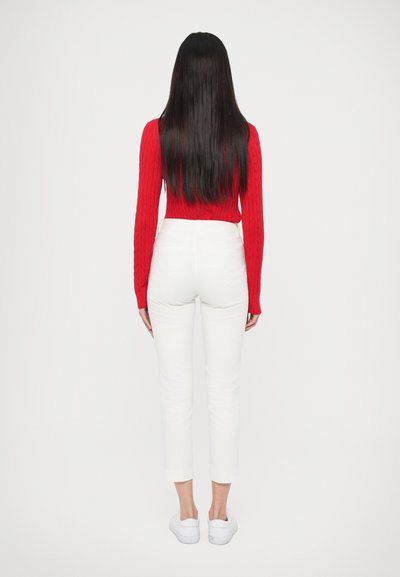 Κόκκινο πλεκτό πουλόβερ με ριγέ λεπτομέρειες και μακριά μανίκια, λευκό παντελόνι στενής γραμμής, συνδυασμένο με λευκά αθλητικά παπούτσια. Η ενδυμασία φαίνεται από πίσω πάνω σε απλό φόντο.