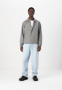 Grå knäppjacka med elastiska ärmar, bärs över en vit t-shirt. Ljusblå vida jeans och kraftiga svarta skor fulländar outfiten.