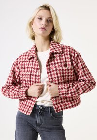 Blonde vrouw die een cropped rood-wit houndstooth jasje over een wit top en donkergrijze spijkerbroek dichtknopt, staande tegen een effen achtergrond.