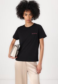 Black cotton t-shirt with "GUESS" logo in red, crew neckline, short sleeves, paired with light-colored satin trousers and a beige clutch bag.