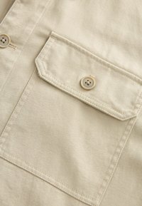 Beige ing szövet, amelyen egy mellzseb található bézs gombbal, egyenes varrással és finom bordázott mintázattal.