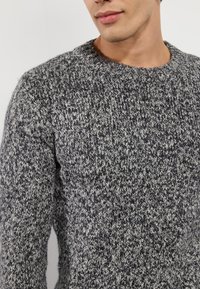 Mann in einem schwarz-weißen melierten Strickpullover mit Rundhalsausschnitt, der den Oberkörper und das untere Gesicht vor einem schlichten Hintergrund zeigt.
