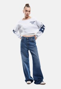 Sudadera recortada de color gris claro con texto y acentos en azul marino; jeans de tiro alto y pierna ancha de color azul; el modelo lleva zapatillas beige.