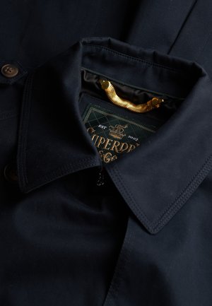 Veste bleu marine avec un col structuré, une fermeture éclair en métal et des accents en bouton marron. Présente une étiquette de marque verte avec des lettres dorées.