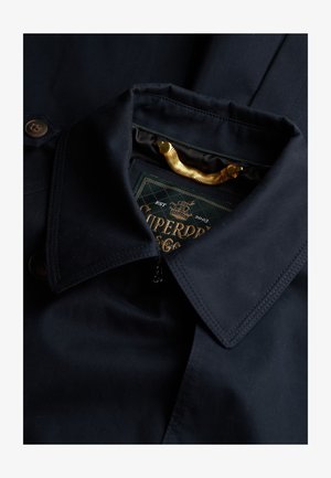 Giacca blu navy con colletto strutturato, zip in metallo e dettagli in bottone marrone. Presenta un'etichetta verde del marchio con lettering dorato.