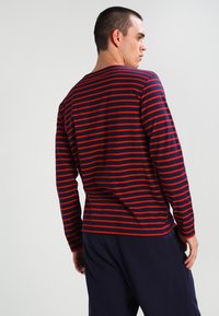 Camicia a maniche lunghe di colore blu navy con strisce rosse orizzontali. Presenta un collo a giro e un tessuto morbido, indossata con pantaloni scuri.