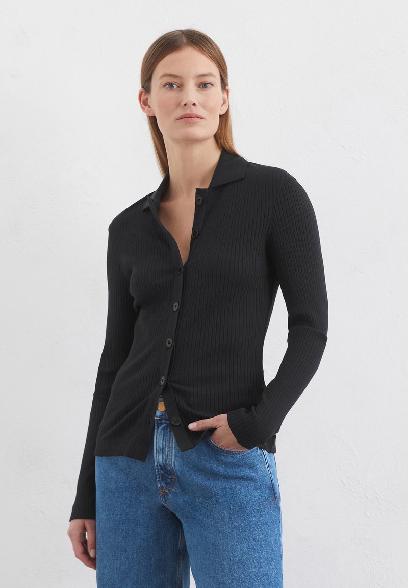 Schwarzes, geripptes Langarm- Hemd mit Knopfverschluss, das einen gekerbten Kragen und eine glatte Textur aufweist, kombiniert mit blauen Jeans.