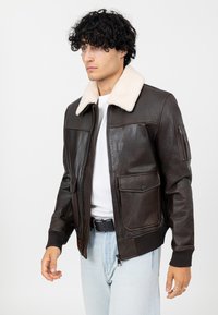 Bruine leren jas met een schapenvacht kraag, ritssluiting aan de voorkant, twee borstzakken en geribbelde manchetten. Gekozen over een wit t-shirt.