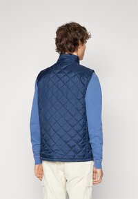 Gepolsterte marineblaue Weste mit hohem Kragen, leichtem Material und seitlichen Taschen, über einem blauen Langarmhemd getragen.