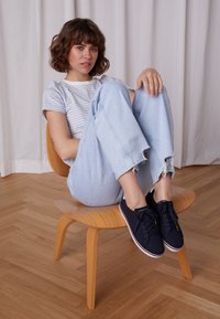 Námořnicky modré plátěné tenisky s bílými tkaničkami, červeným a bílým lemem na podrážce, nošené s světle modrými džíny s potrhaným efektem a proužkovaným tričkem, sedící na dřevěné židli.