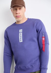 Felpa viola con collo a giro, dotata di un'etichetta grigia "ALPHA" e una tasca con zip rossa sulla manica. Tessuto in cotone testurizzato.