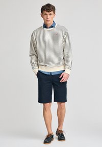 Pasiasty sweter w kolorze kremowym i granatowym założony na niebieską koszulę dżinsową, w komplecie z ciemnonawowymi szortami i granatowymi butami z brązowymi sznurówkami.