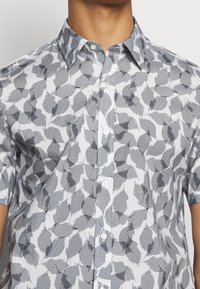 Lyhythihainen paita valkoisesta kankaasta, jossa on harmaa lehtikuosi, kaulus ja napitettava edusta. Sileä pinta ja graafinen muotoilu.