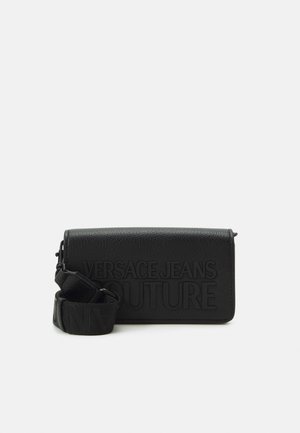 Zwarte leren Versace Jeans Couture crossbodytas met reliëflogo en brede verstelbare schouderband, opgerold naast de tas op een witte achtergrond.