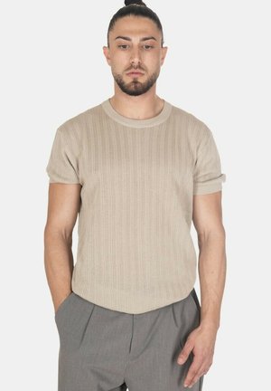Uomo con barba e capelli raccolti indietro che indossa una maglia beige a maniche corte e pantaloni grigi, in piedi con le mani nelle tasche.
