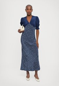 Donkerblauwe jurk met korte pofmouwen, stippenpatroon en een uitlopende zoom. Ggedragen met een crèmekleurige clutch en witte puntige hakken.