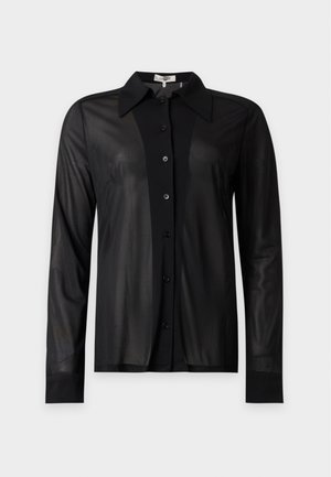 Femme mince portant une chemise noire transparente à manches longues boutonnée, un pantalon rose vif et des talons hauts ouverts noirs, debout devant un fond uni.
