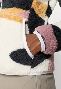 Fleece jaka ar abstraktu rakstu melnā, rozā, zelta un baltā krāsā. Iezīmēta ar baltu apdari ap piedurknēm un kabatām. Mīksts, teksturēts audums.