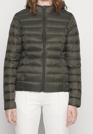 Femme portant une doudoune matelassée vert olive avec fermeture éclair à l'avant et pantalon blanc, se tenant contre un fond clair uni.