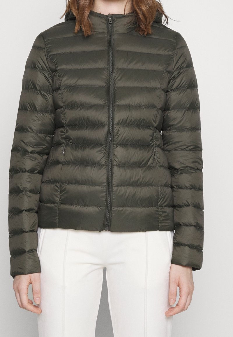 Femme portant une doudoune matelassée vert olive avec fermeture éclair à l'avant et pantalon blanc, se tenant contre un fond clair uni.