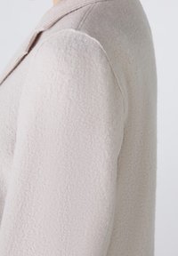 Abrigo de lana beige claro con un acabado texturizado, que presenta un cuello con solapa y un corte entallado. La vista lateral resalta la costura del hombro.