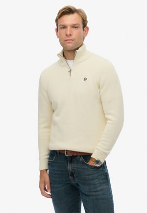 Pull zipé couleur crème avec col et poignets côtelés, arborant un petit logo brodé sur la poitrine. Porté avec un jean bleu et une montre.