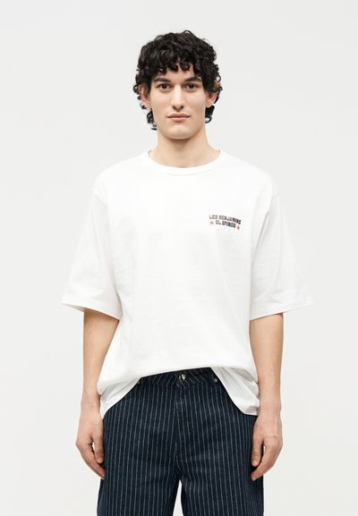 Νέος άνδρας με σγουρά μαλλιά, φοράει λευκό φαρδύ T-shirt με μικρό κόκκινο κείμενο και μαύρο παντελόνι με λεπτές ρίγες, στέκεται μπροστά σε λευκό φόντο.